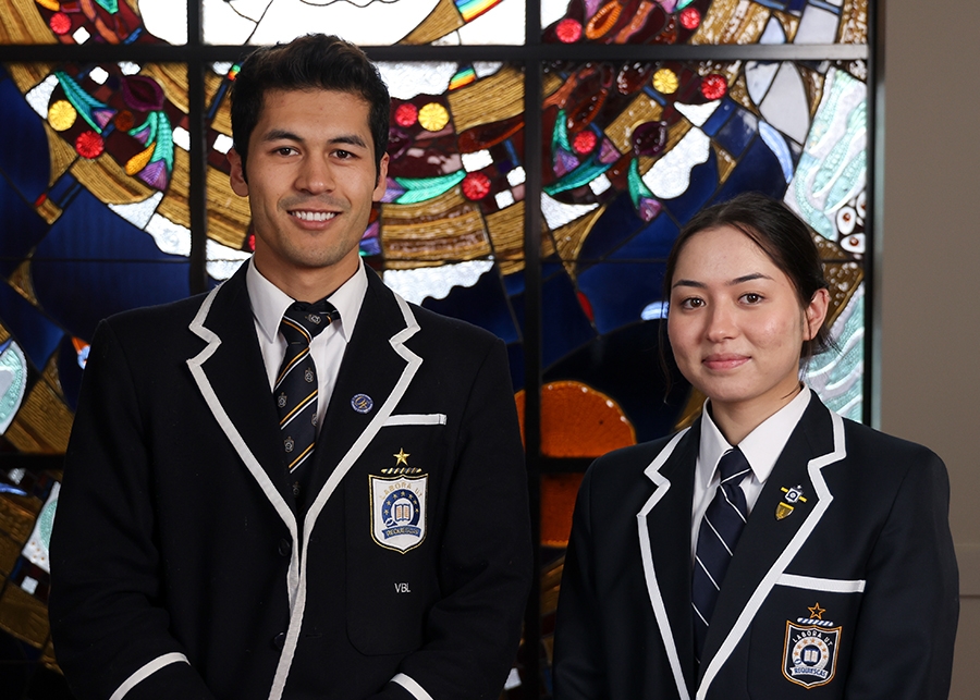 Two students from Caulfield Grammar School in uniform pose in front of a colourful stained-glass window.